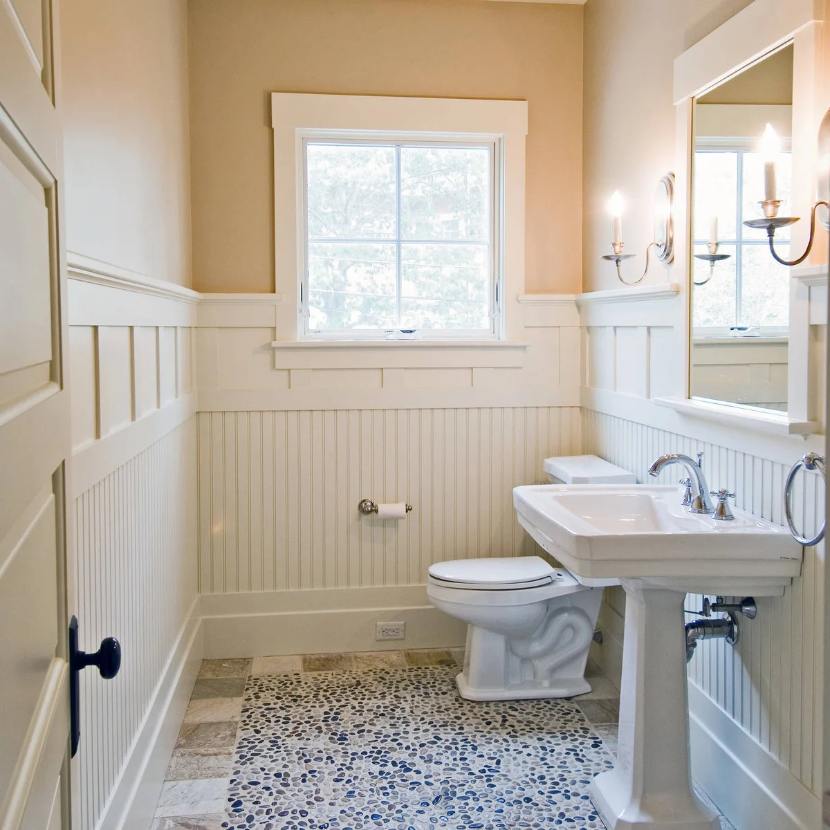 A white pedestal sink and toilet sit in a pale room with beadboard, wider upper panels, a window and a pebble-patterned floor inset.