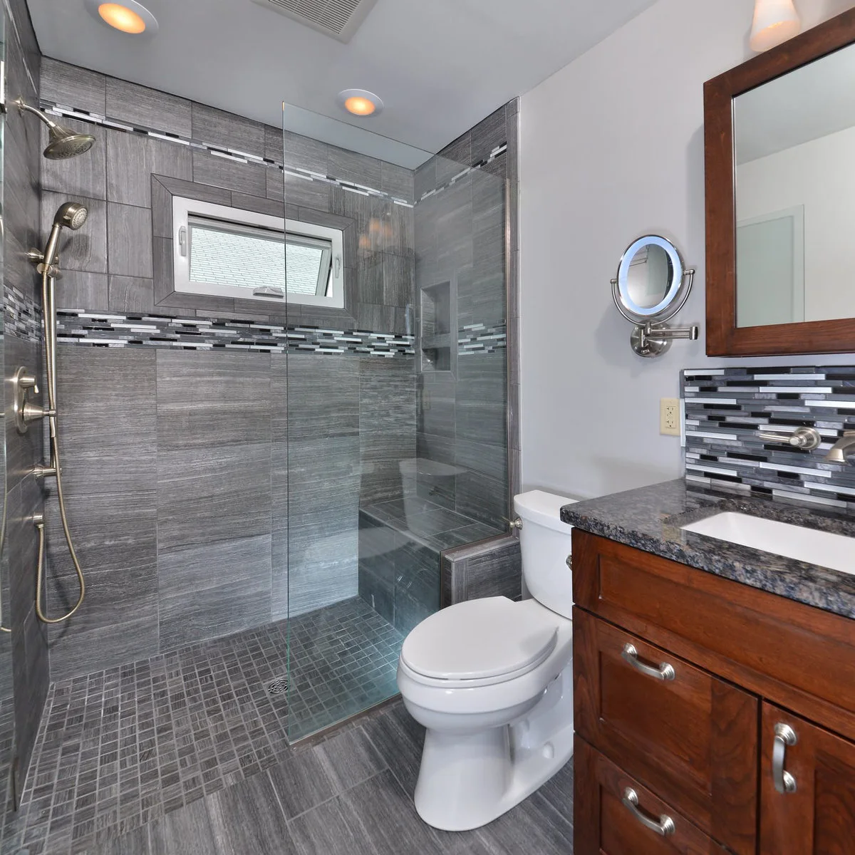 Gray tiled shower with glass above a short wall beside a toilet and wood vanity