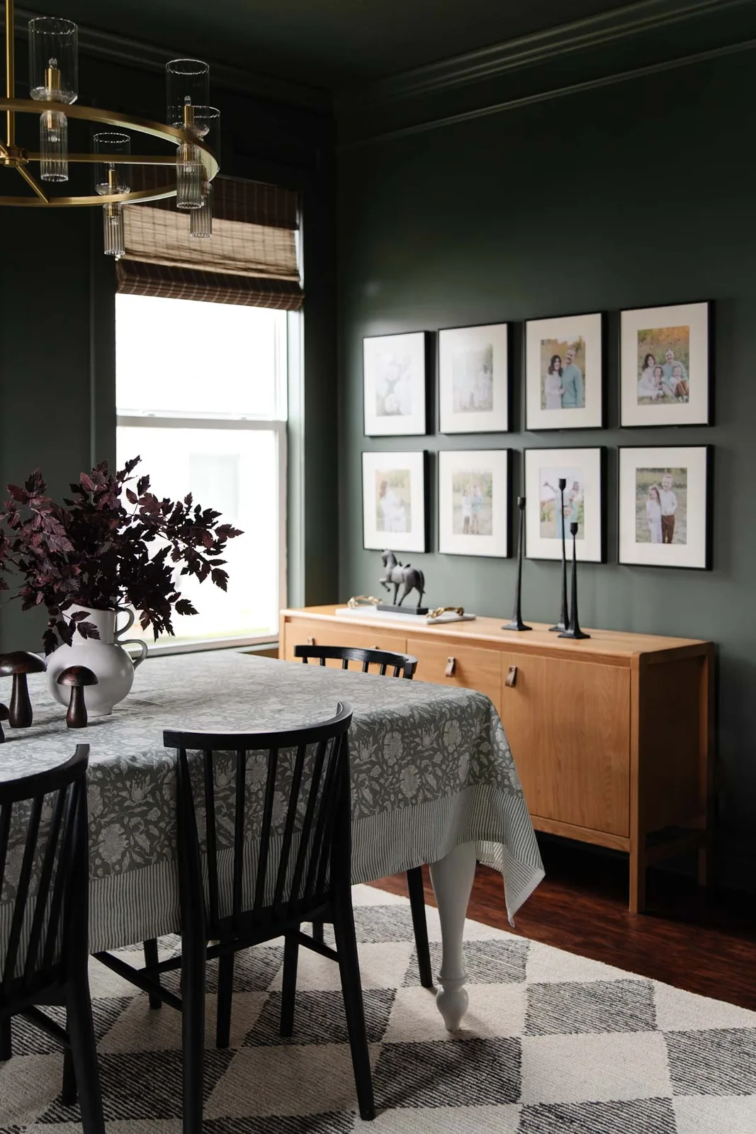 A warm wood-toned sideboard sits beneath eight framed photographs beside a deep green dining wall, black chairs and pale patterned tablecloth