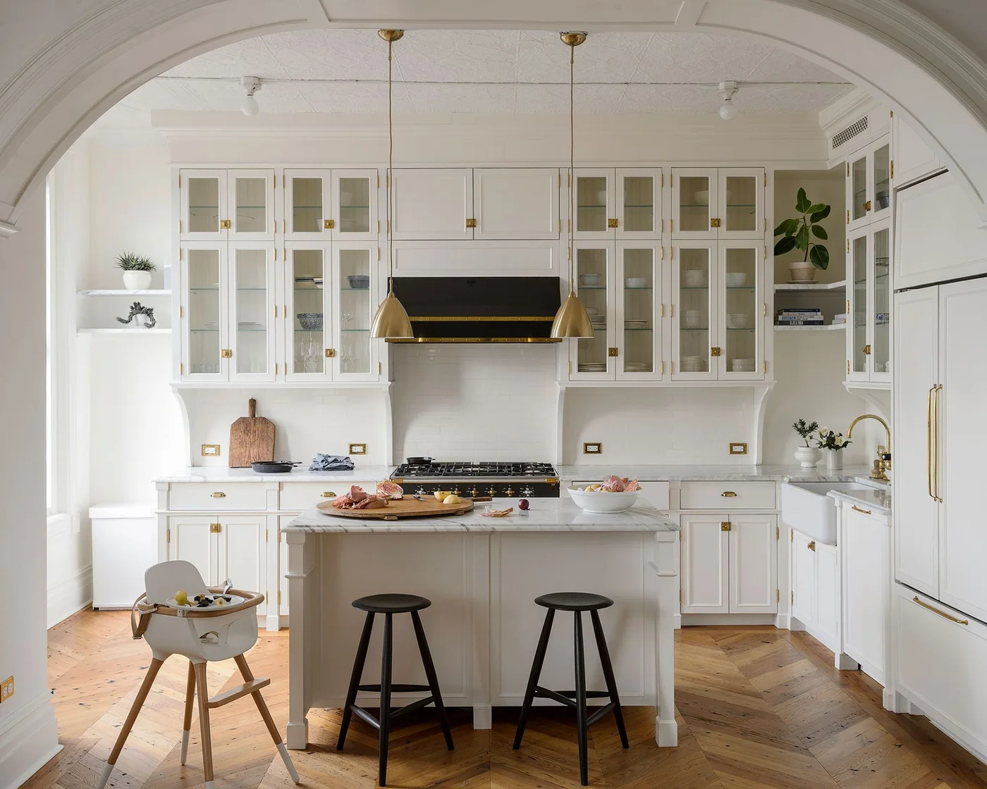 White glass-front cupboards surround a small paneled island beneath slim warm metal pendants and an arched opening.