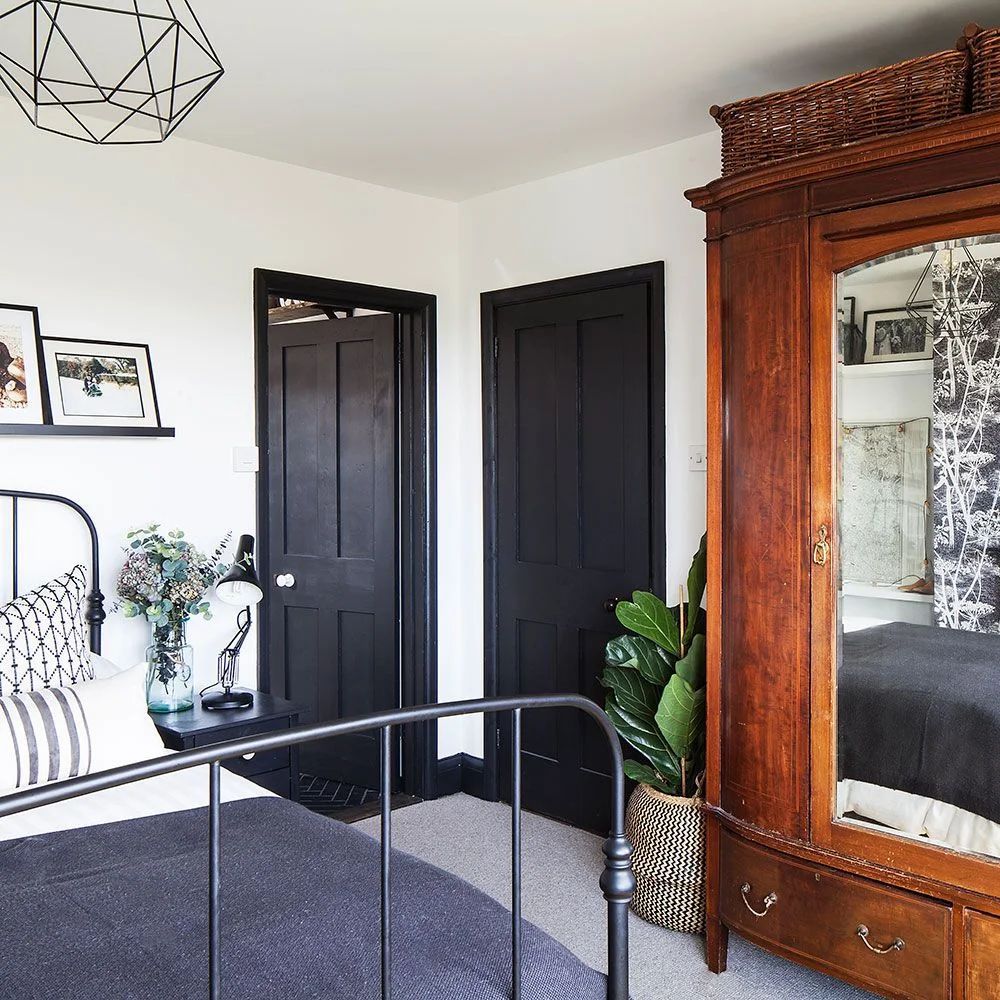 A dark open bed frame sits beside black doors, white walls and a warm wood wardrobe with a mirrored door and baskets above