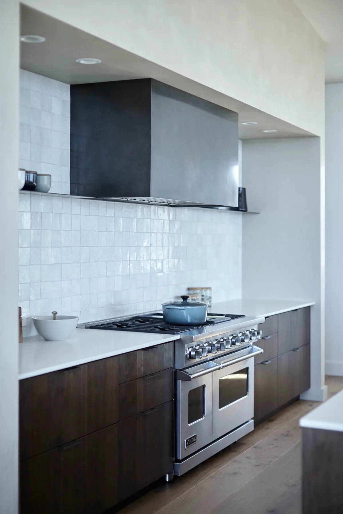 Dark brown lower kitchen cabinets beneath pale counters, white square tiles and a broad metal hood.
