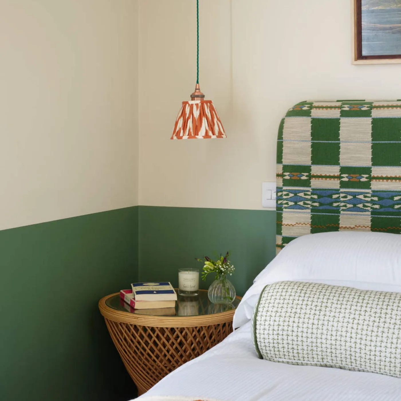 A deep green lower wall sits beneath pale paint beside a green checked headboard, woven bedside table and orange-patterned pendant