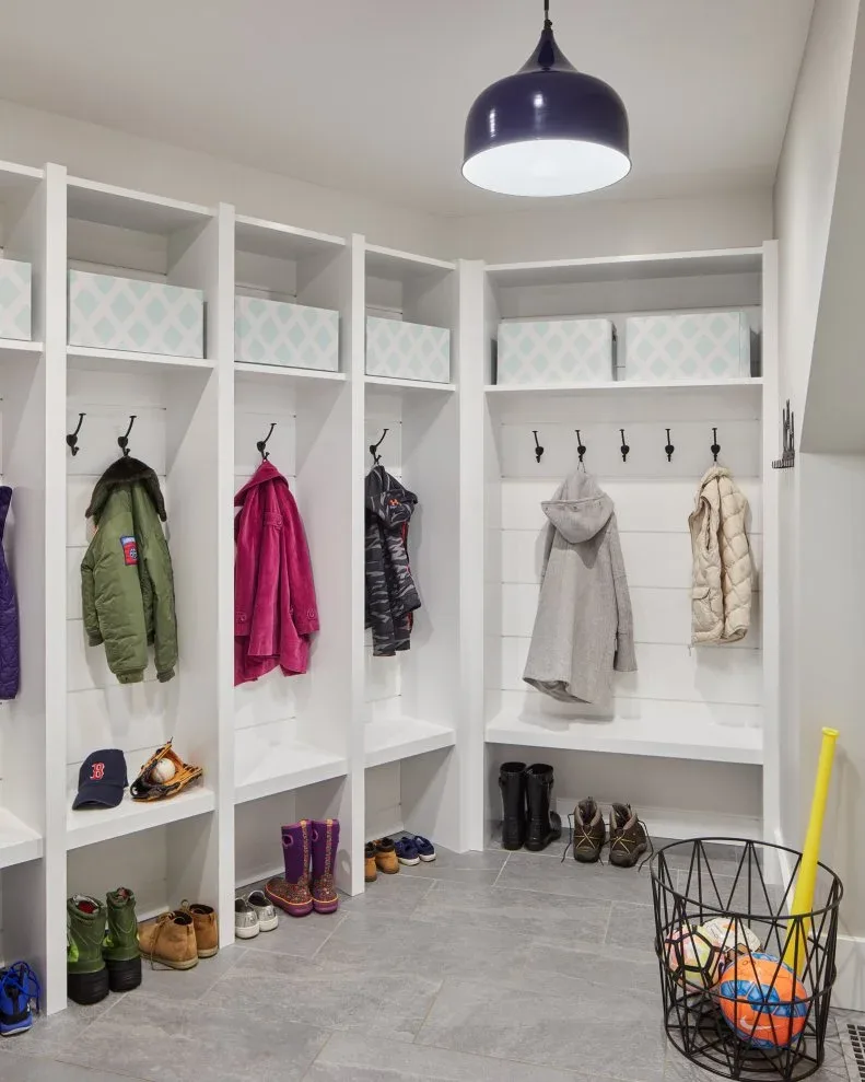 White bench seating turns a corner beneath coat hooks and open cubbies, with boots stored below and a sports basket nearby.