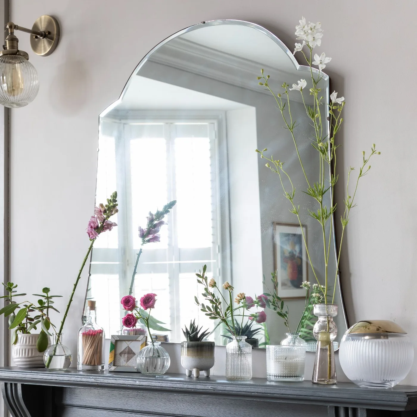 Dark mantel with arched mirror, varied small glass vases, individual flowers and pale ribbed container