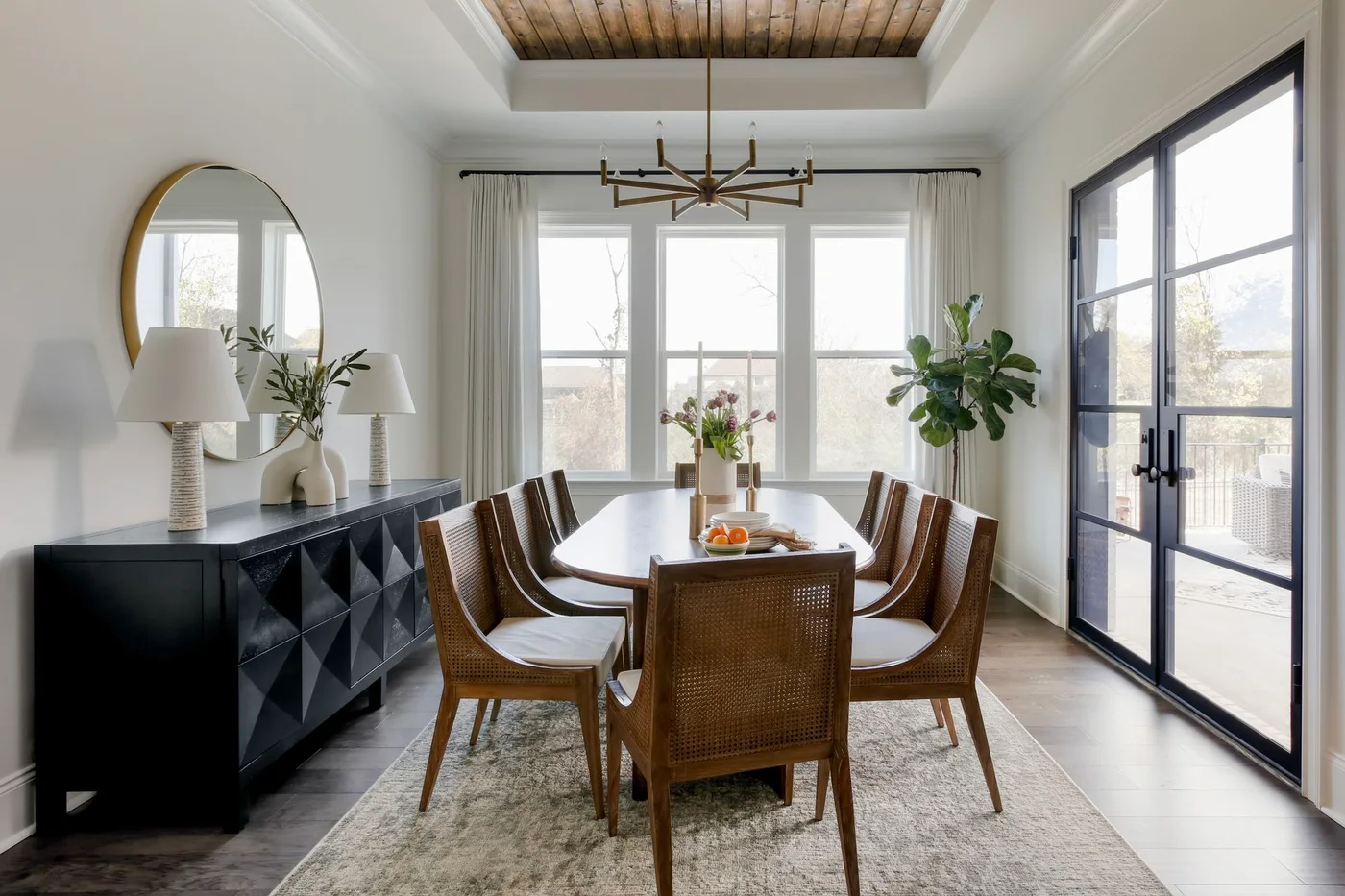 A dark sideboard with geometric faceted fronts stands beneath a round mirror and two pale lamps beside a wood dining table and woven-back chairs