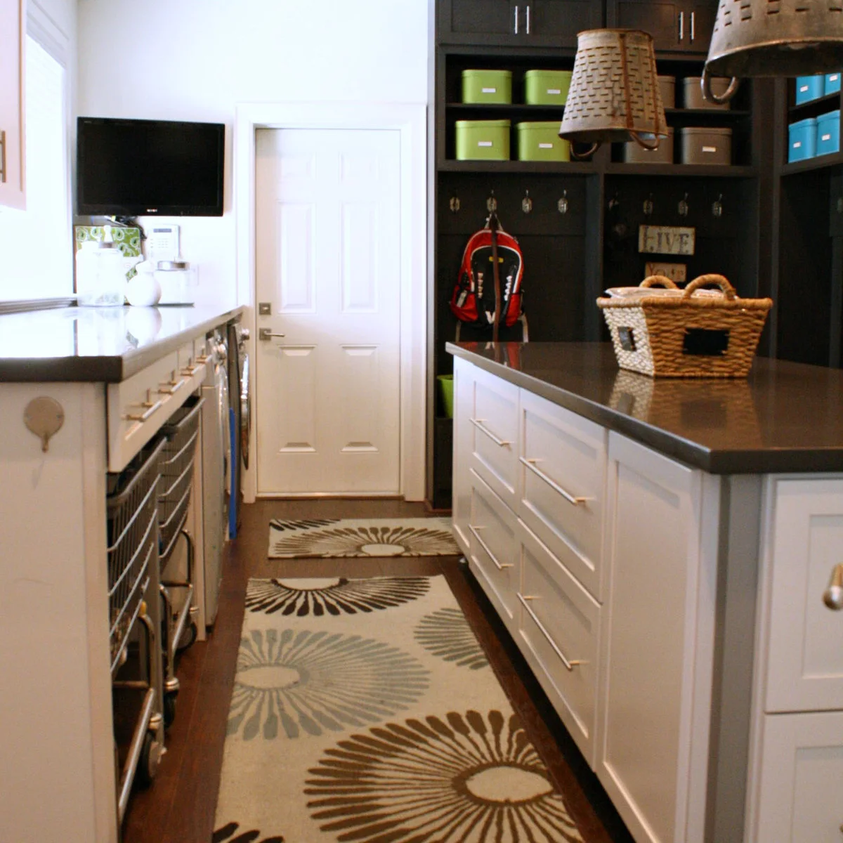 Large white laundry room with an island, appliance counter and a dark coat-and-basket storage wall at the back