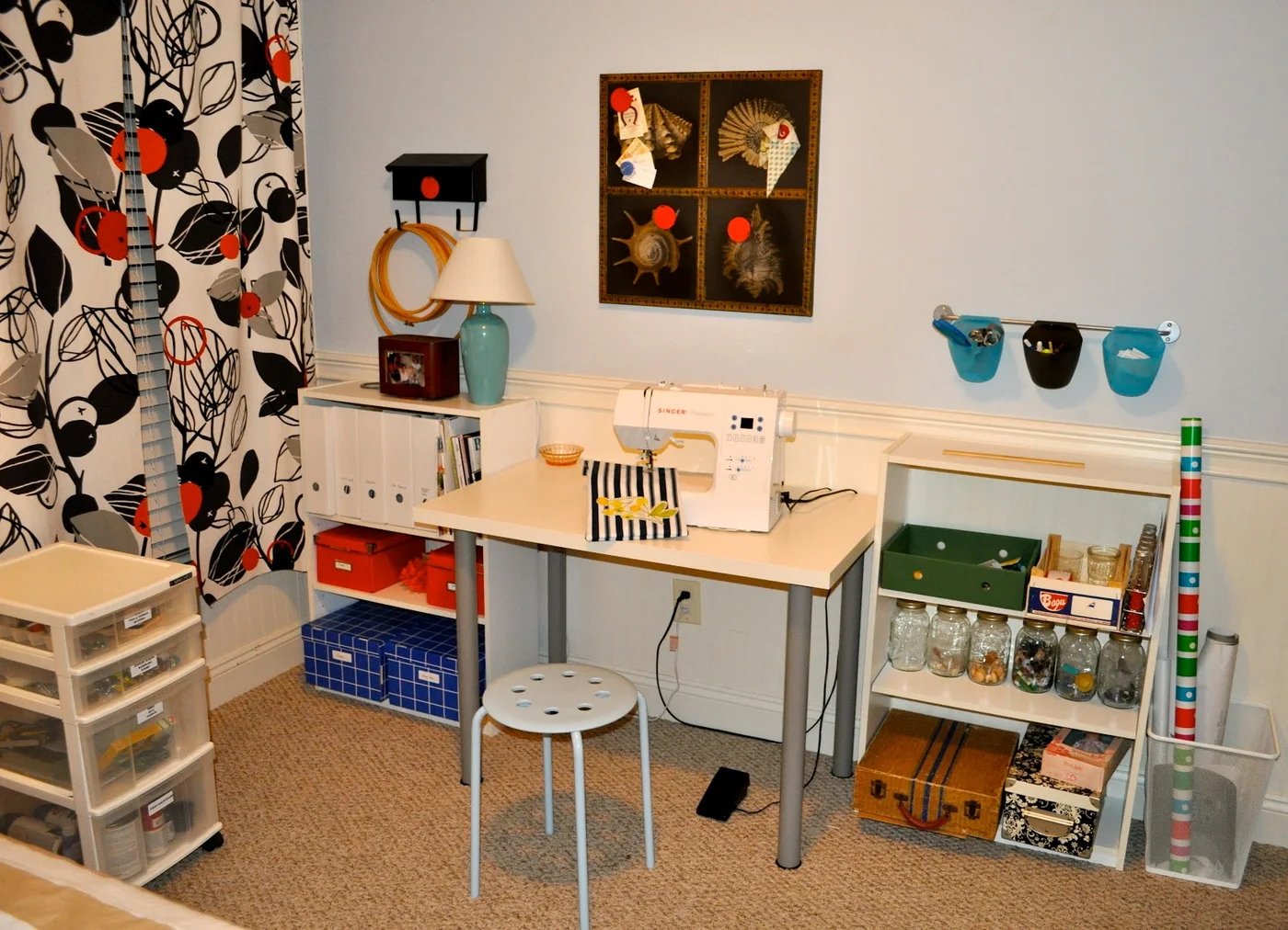 A sewing machine on a pale desk sits between short shelves, with a small stool, wall-hung cups and a rolling clear drawer unit nearby.