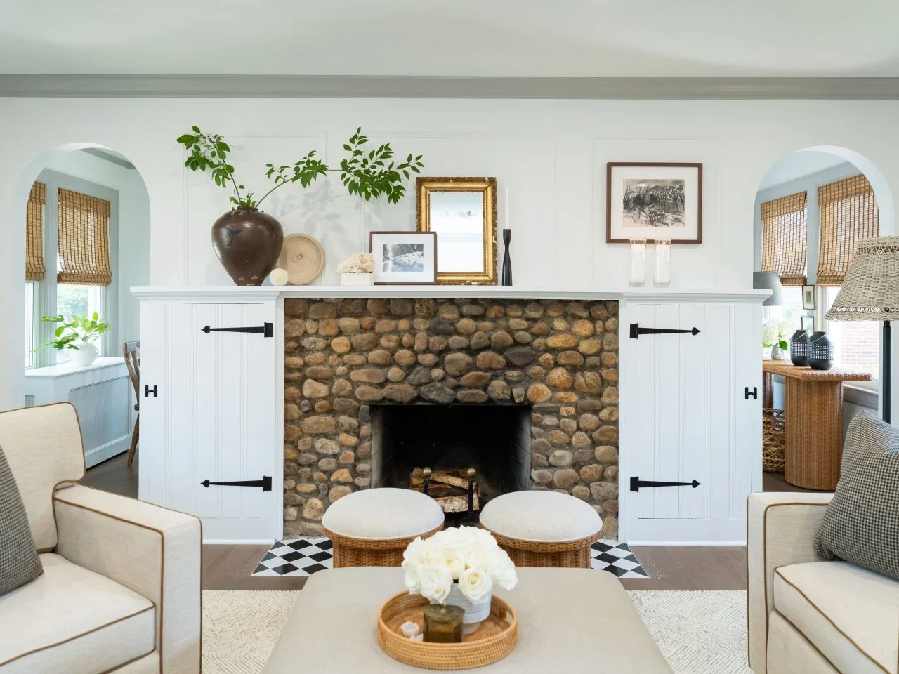 Pale armchairs and woven stools face a rounded-stone fireplace between white paneled cupboards.