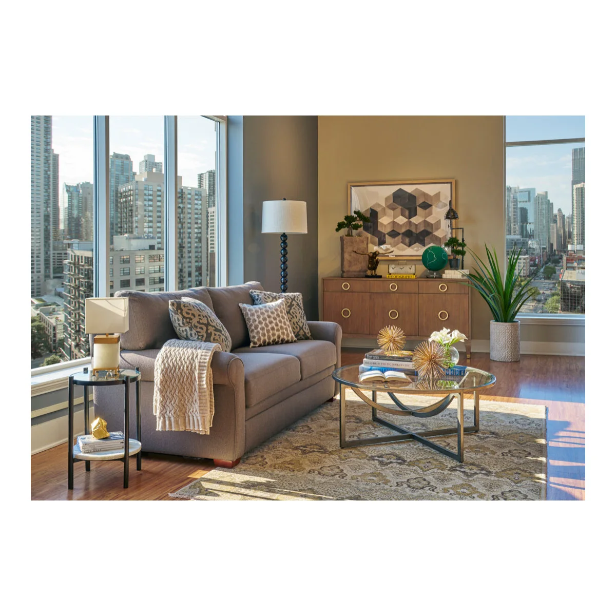Taupe sleeper sofa beside tall windows with a round glass coffee table and a small side table
