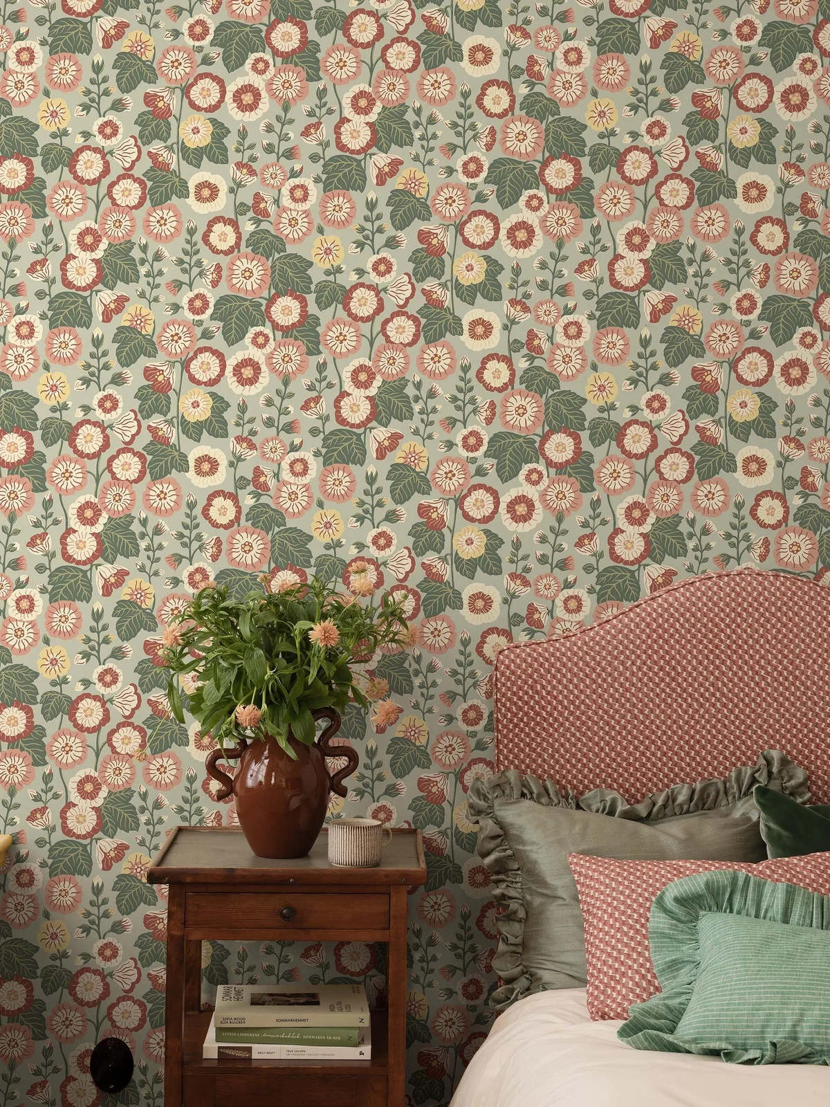Red and cream flowers with green leaves repeat behind a red patterned upholstered headboard, green cushions and a small older wood bedside table.