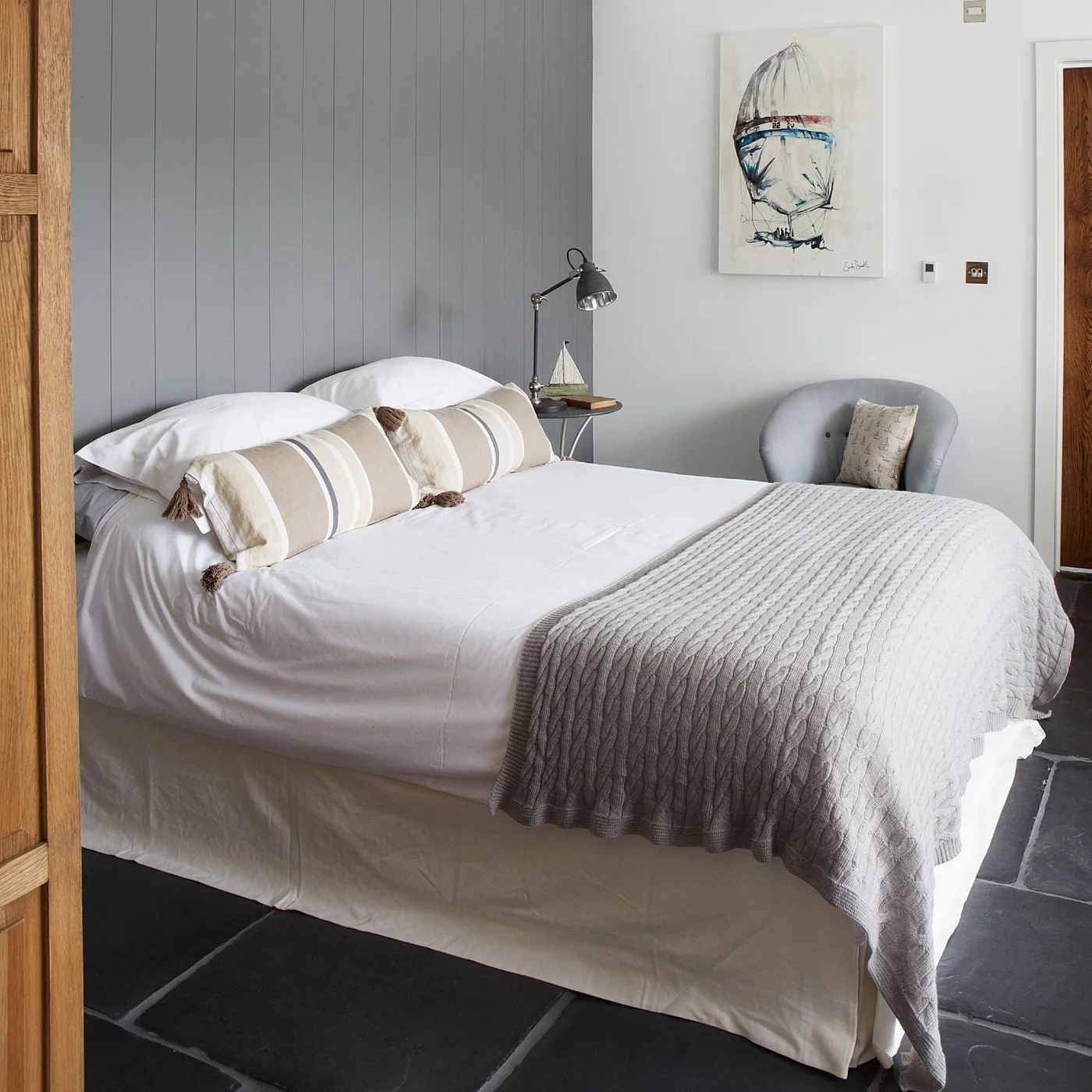 A white bed with a pleated valance and gray knitted throw stands against a gray vertical-board wall with a small round table and an upholstered chair.