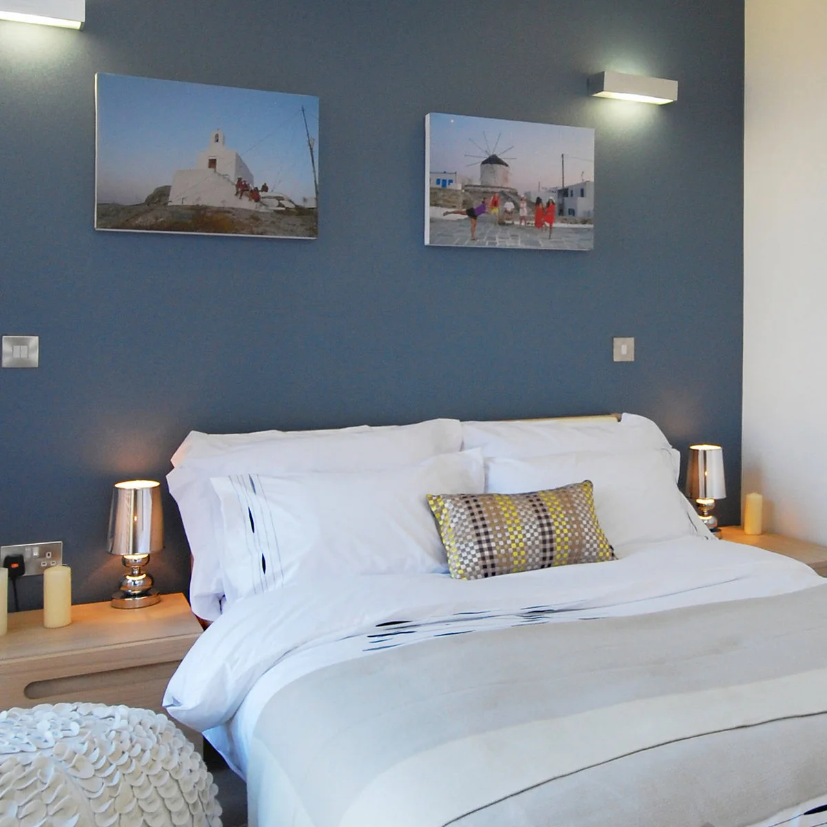 Slate blue bedroom wall with two travel photographs, white bedding and small metallic bedside lamps.