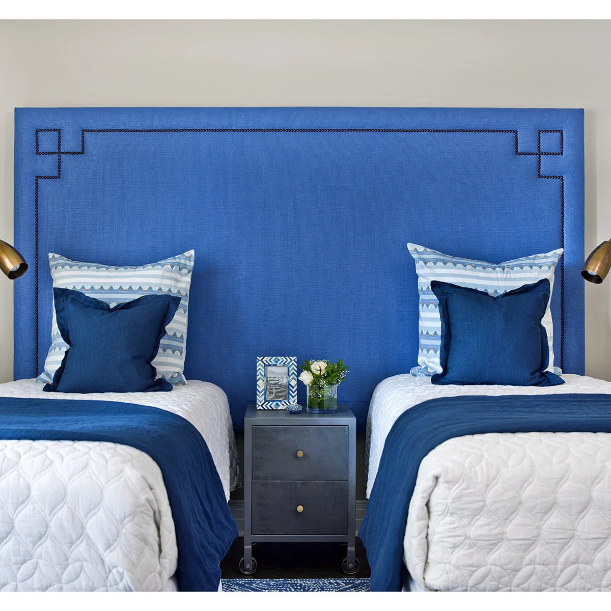 Twin beds with white quilts and blue throws beneath a shared blue upholstered headboard.