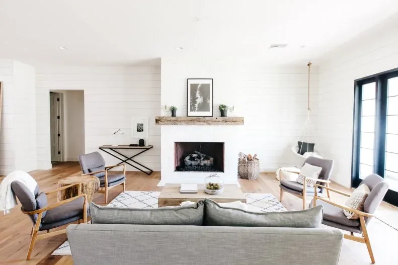 White board-walled living room with pale sofa, wood-framed chairs, simple wood mantel, patterned rug and black-framed glazed doors