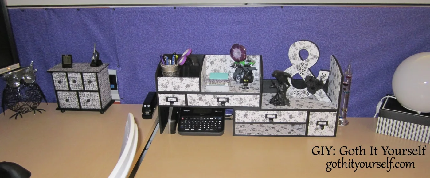 Black and white patterned desktop drawers and open organizers against purple cubicle fabric with a pen cup and label maker below.