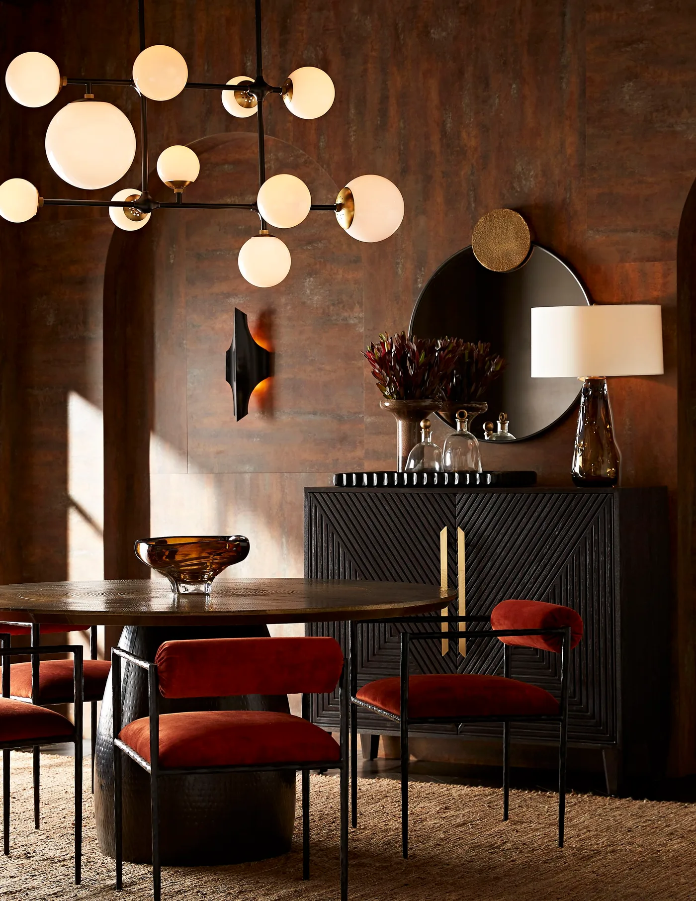 Black sideboard with diagonal ribbing and gold handles beneath a round mirror beside red dining chairs