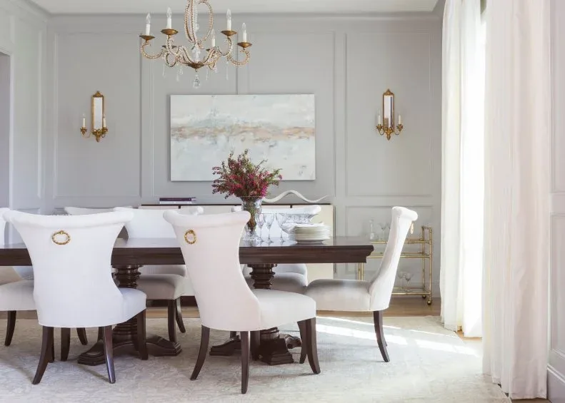 Pale gray rectangular wall frames behind white upholstered dining chairs, a dark table and gold-toned sconces