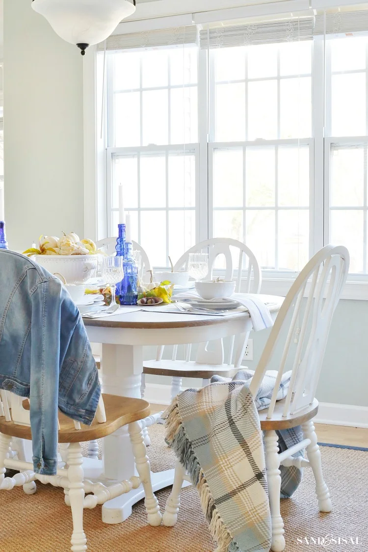 A round wood dining table with white legs and chairs holds white pumpkins, yellow gourds and blue candle bottles beside broad windows