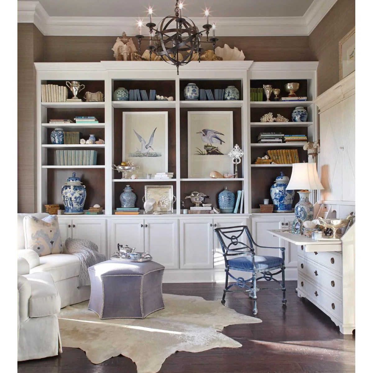 A white secretary desk beside pale armchairs and a wall of bookshelves with blue-and-white ceramics.