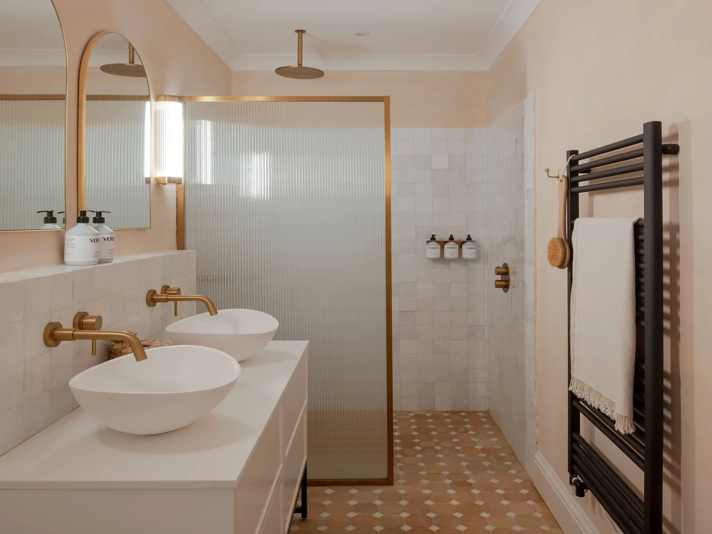 Two oval vessel basins sit beneath warm-metal wall taps beside a ribbed-glass shower enclosure, white square tiles and a muted peach patterned floor.