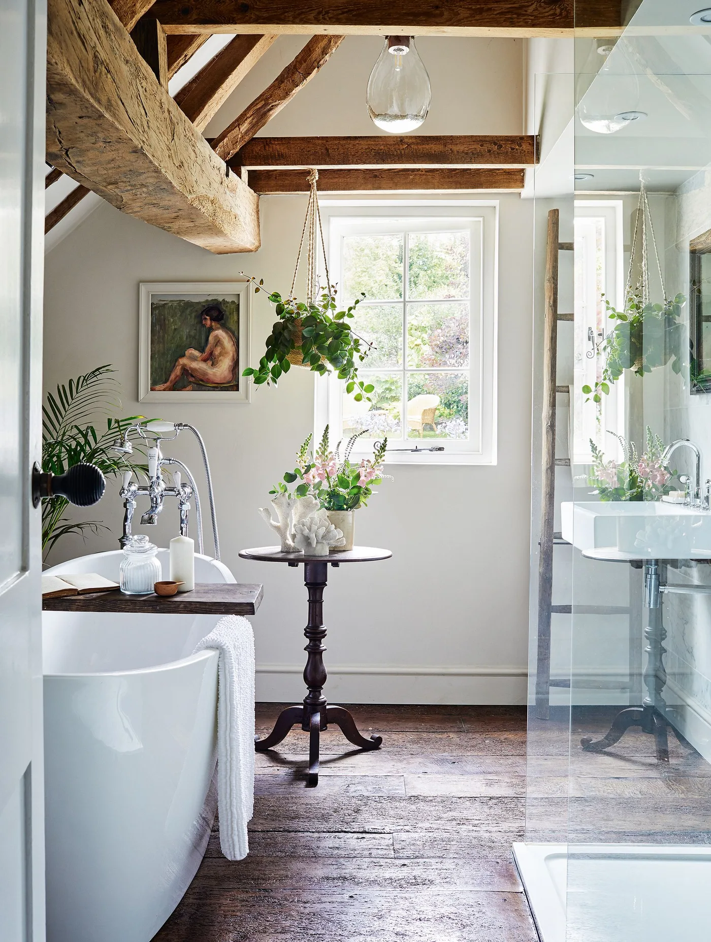 Bathroom with exposed wooden beams, a white bath, hanging plants, wooden floor and a clear shower screen.