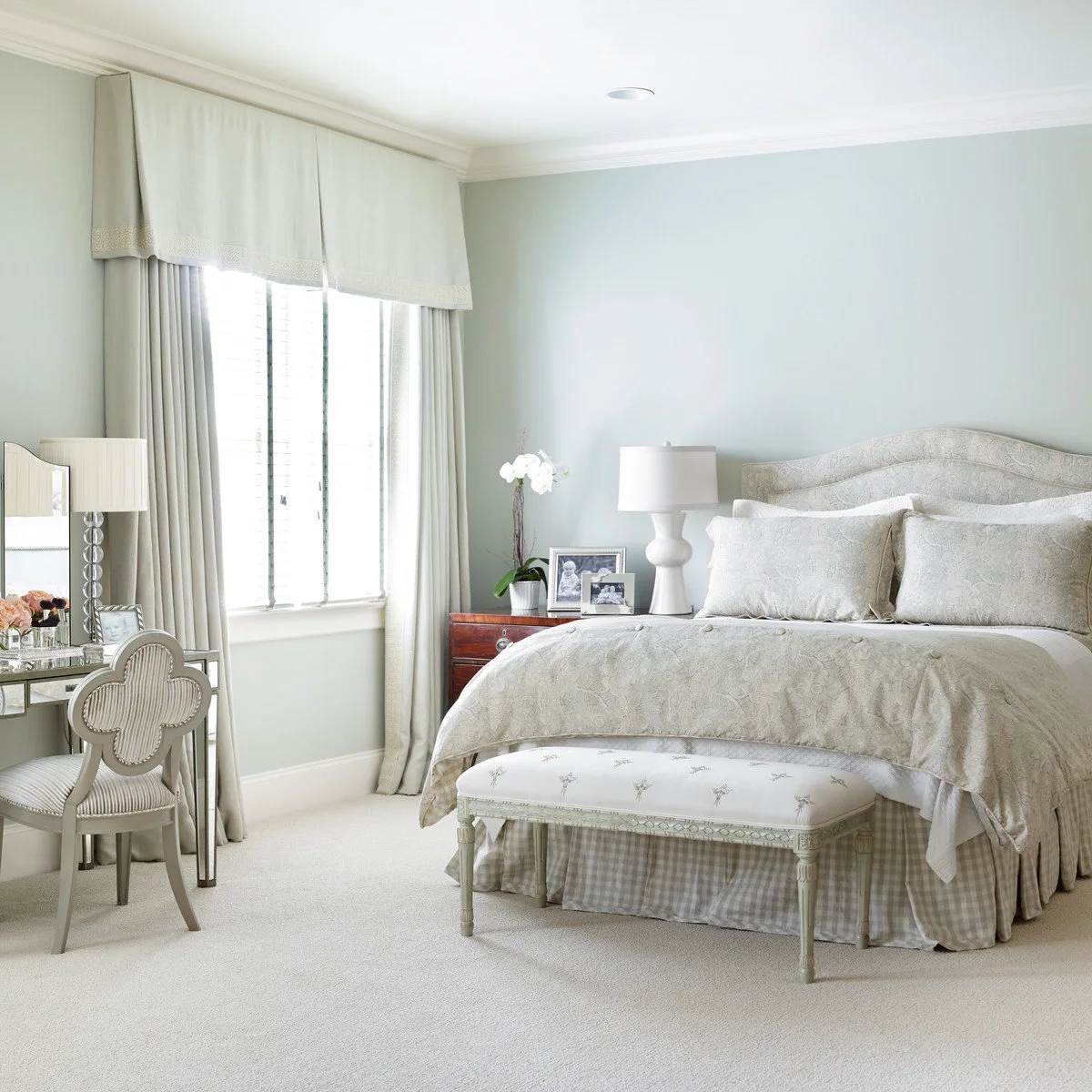 A mirrored dressing table and pale decorative chair sit beside a bed in a softly colored bedroom