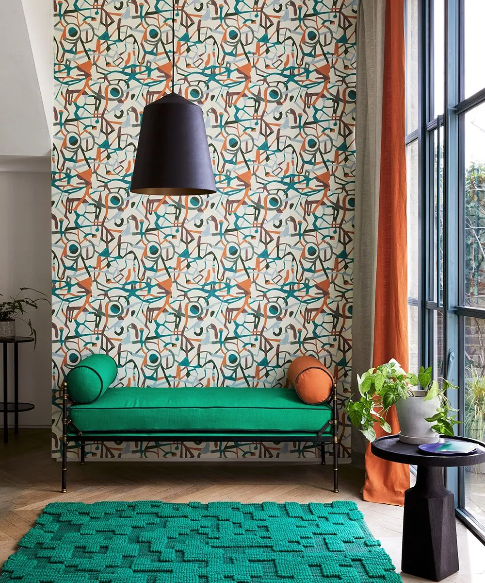 Sitting corner with multicolored abstract wallpaper, solid green chaise, orange bolster, green textured rug, dark pendant and small side table.