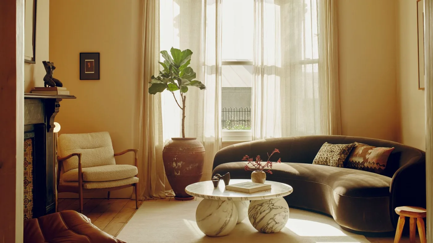 Curved dark brown sofa in a pink and ochre room with a pale round table, white curtains and a potted tree.