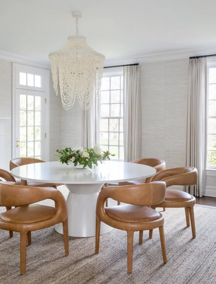 Pale dining room with round white pedestal table, tan curved chairs, light curtains and a fringed chandelier.