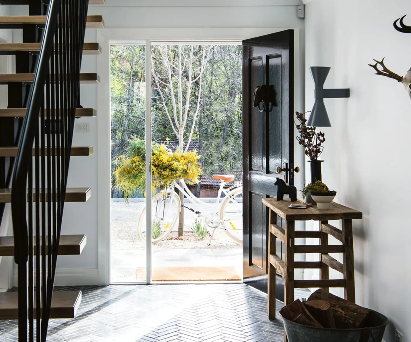 Modern country entry with dark open front door, slim wood console, black stair rails, gray herringbone floor and yellow flowers