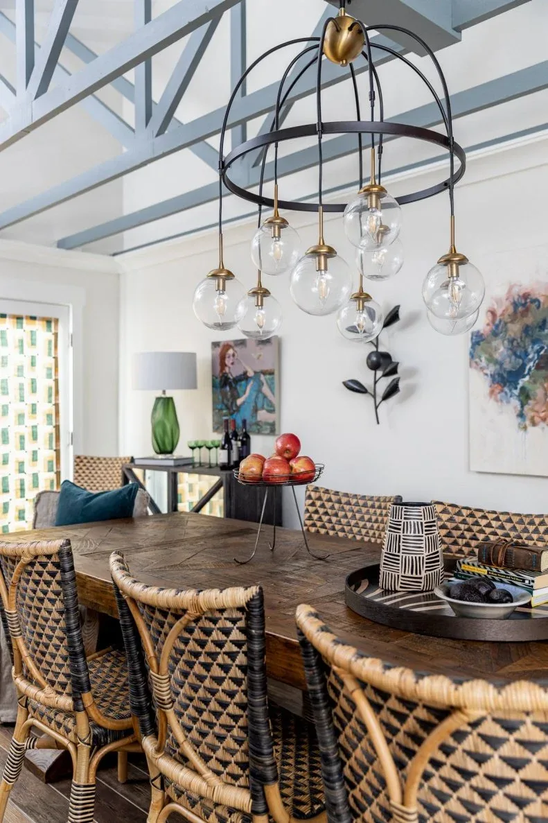 Dark open cage chandelier with clear hanging globes and gold-toned fittings above a wooden dining table and patterned woven chairs, beneath blue ceiling beams.