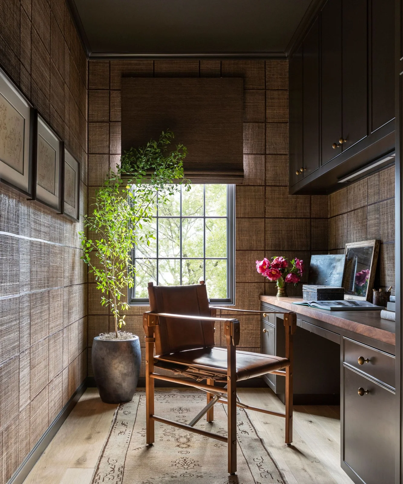 A narrow office has brown textured walls and a matching blind, dark upper cabinets, a wood desk surface and a brown chair beside a window.