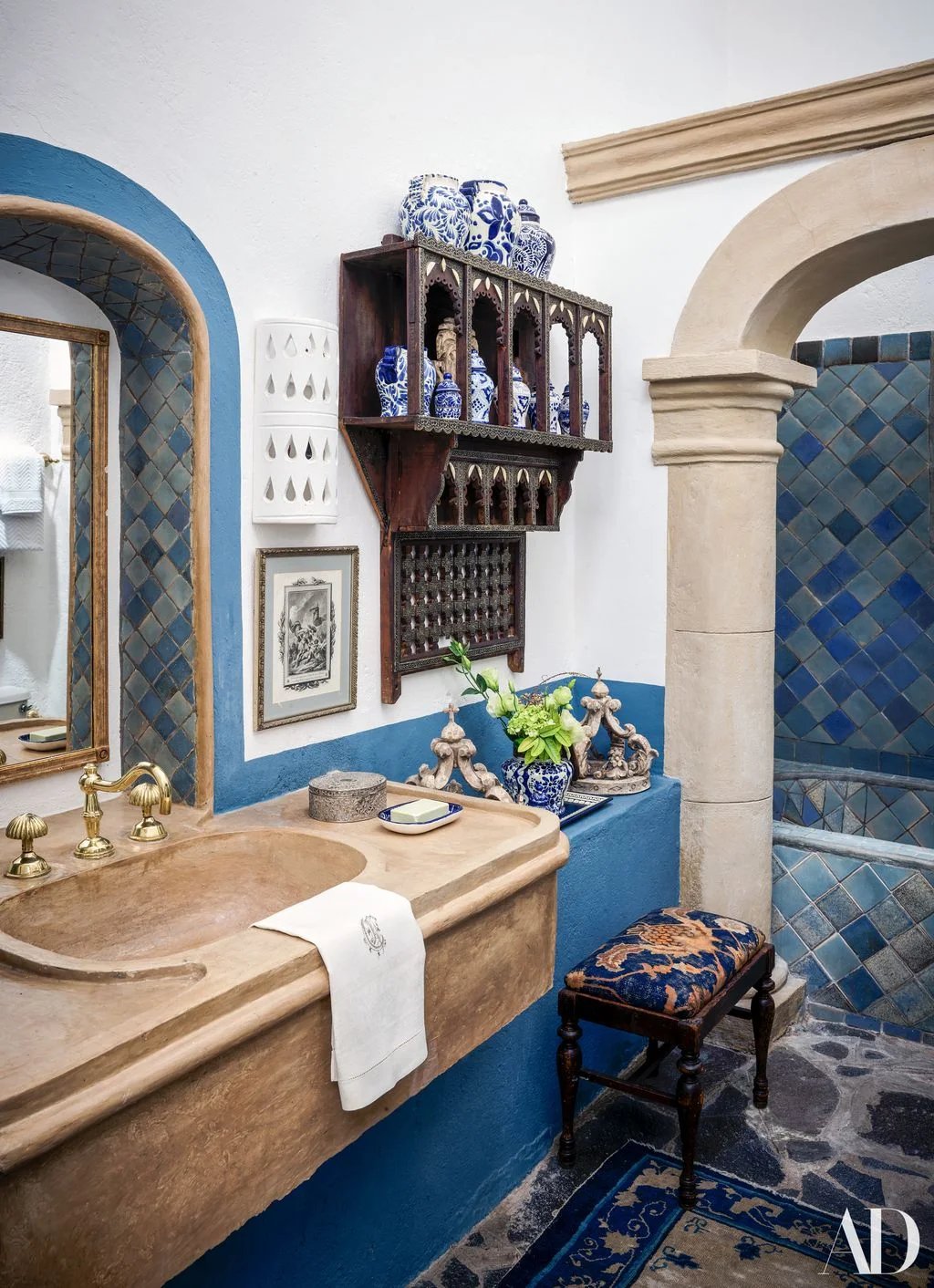 Blue and white pottery on a carved wood wall rack in a bathroom with blue tile and a pale shaped basin surround.