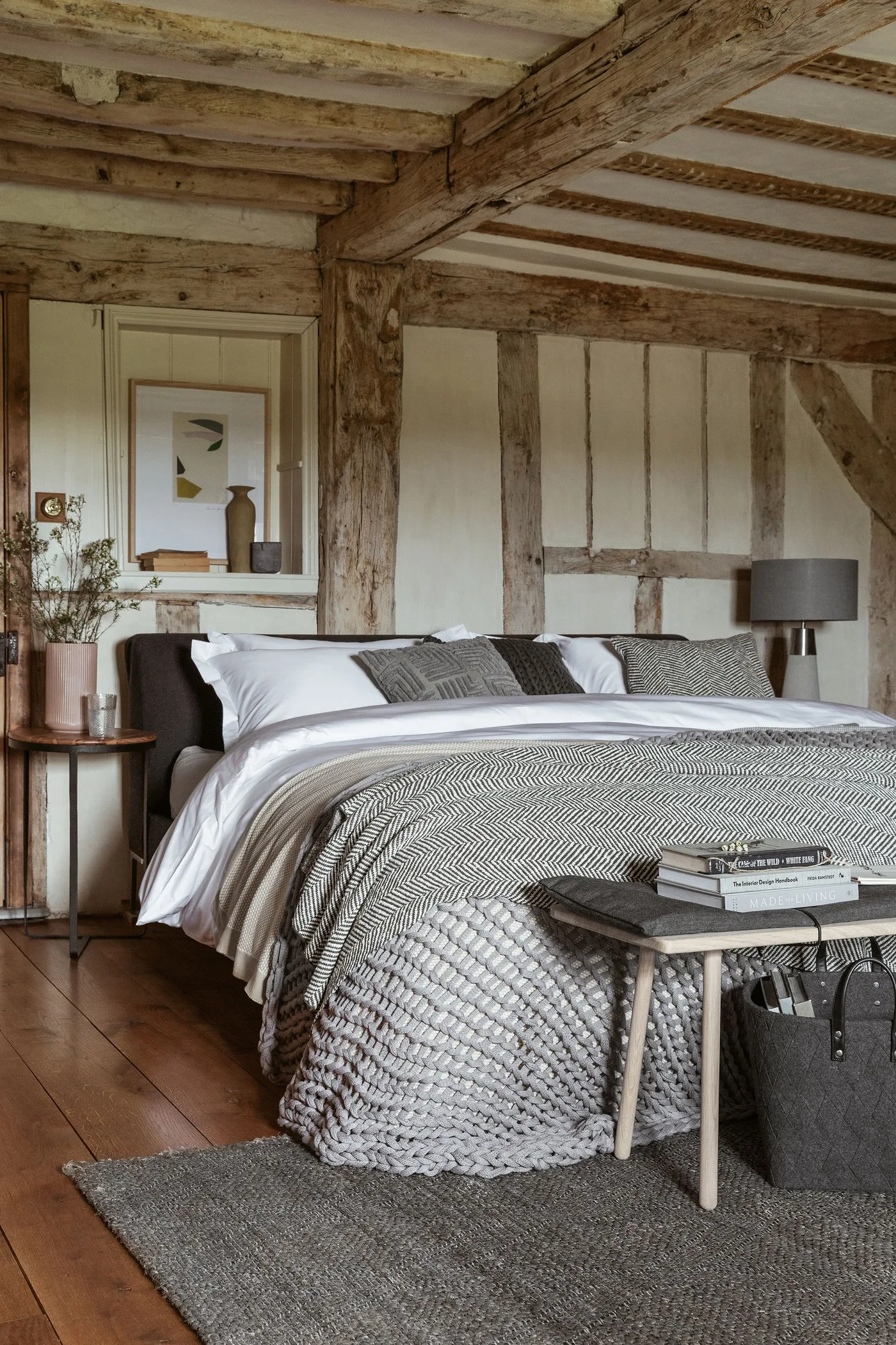 A bedroom with rough timber framing has pale bedding, gray woven layers and a low upholstered headboard.