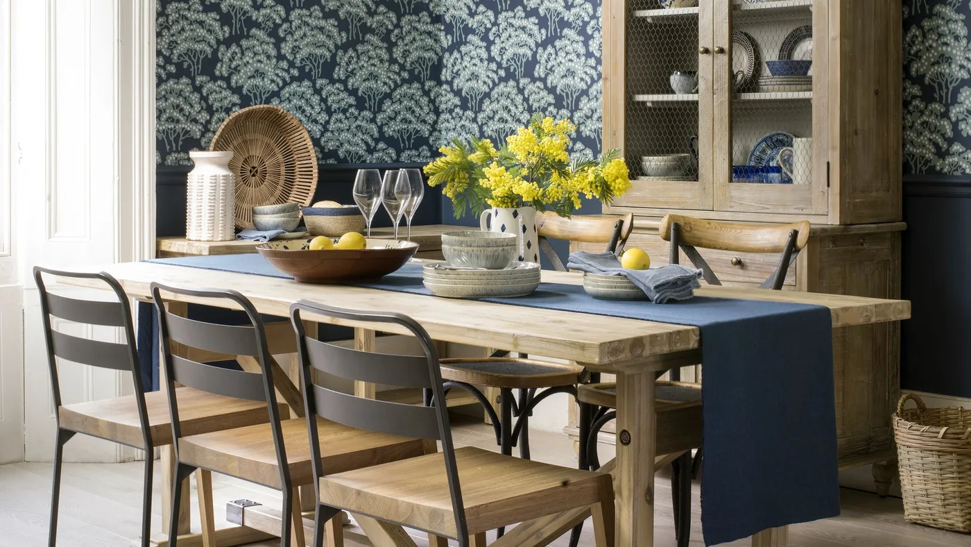 Country dining room with dark blue leafy wallpaper, wooden table, tall wooden dish cupboard, blue runner and mixed dark-framed chairs.
