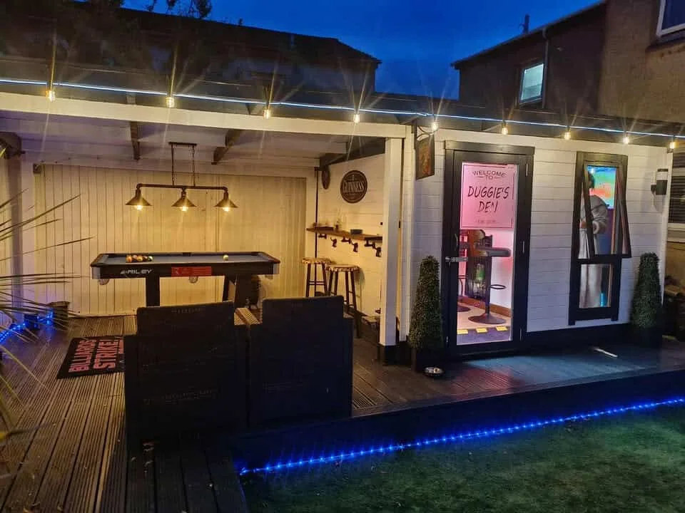 Garden bar cabin beside a covered pool-table area with deck seating and evening lights
