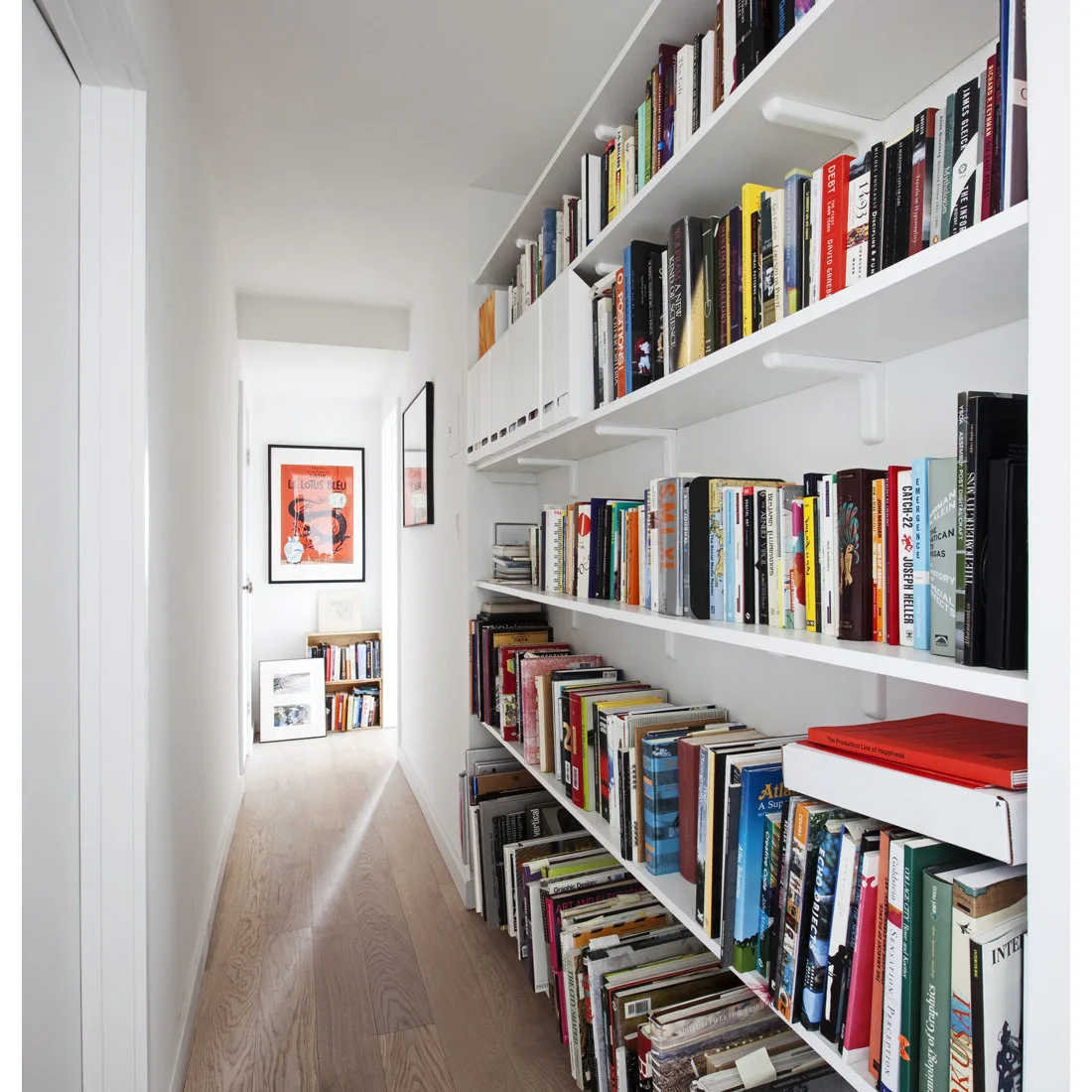 White open shelves filled with books run along one wall of a narrow pale hallway toward framed artwork.