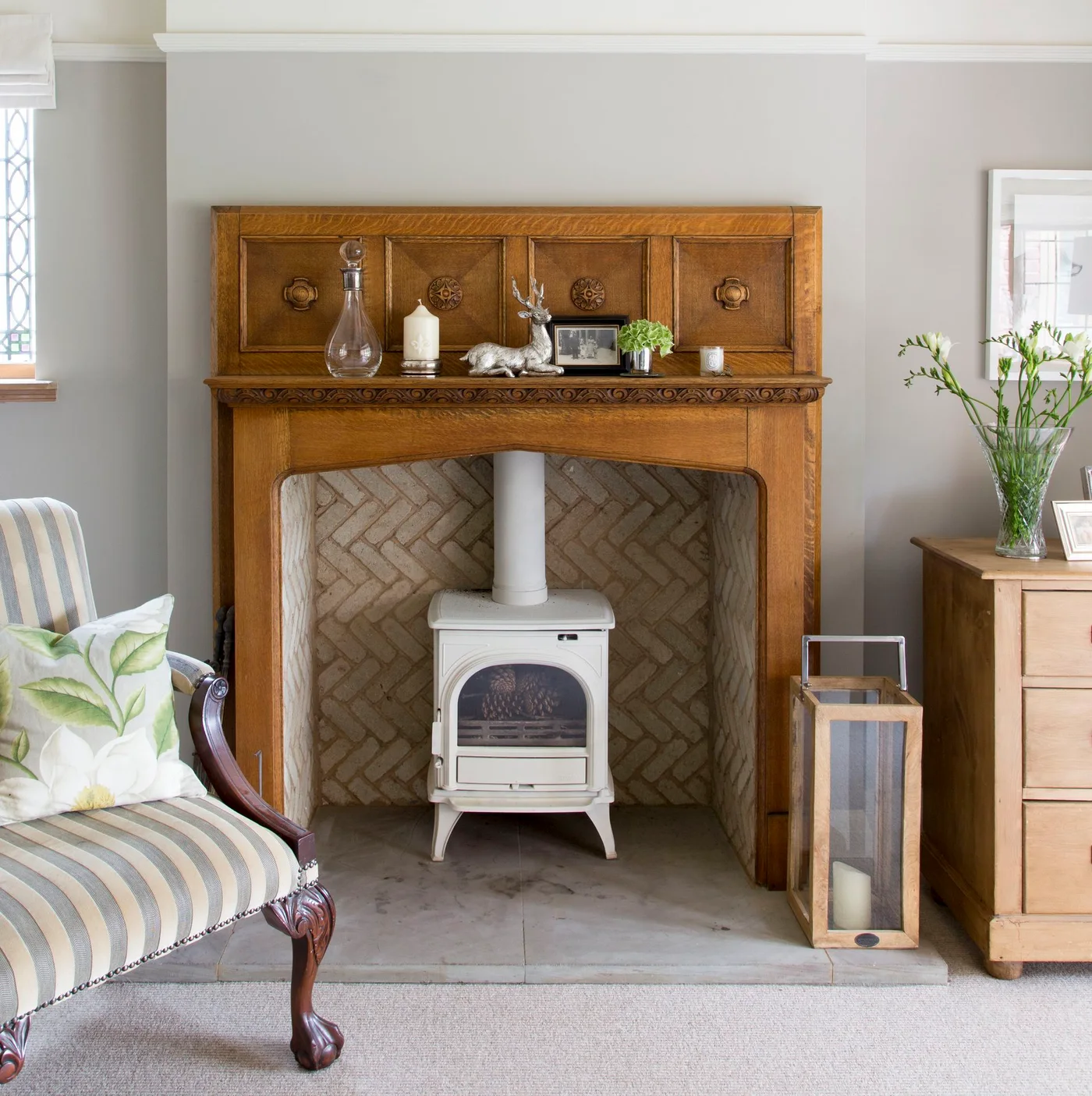 A pale herringbone lining sits behind a small white stove within a carved wood fireplace surround, with a pale hearth and striped chair nearby.