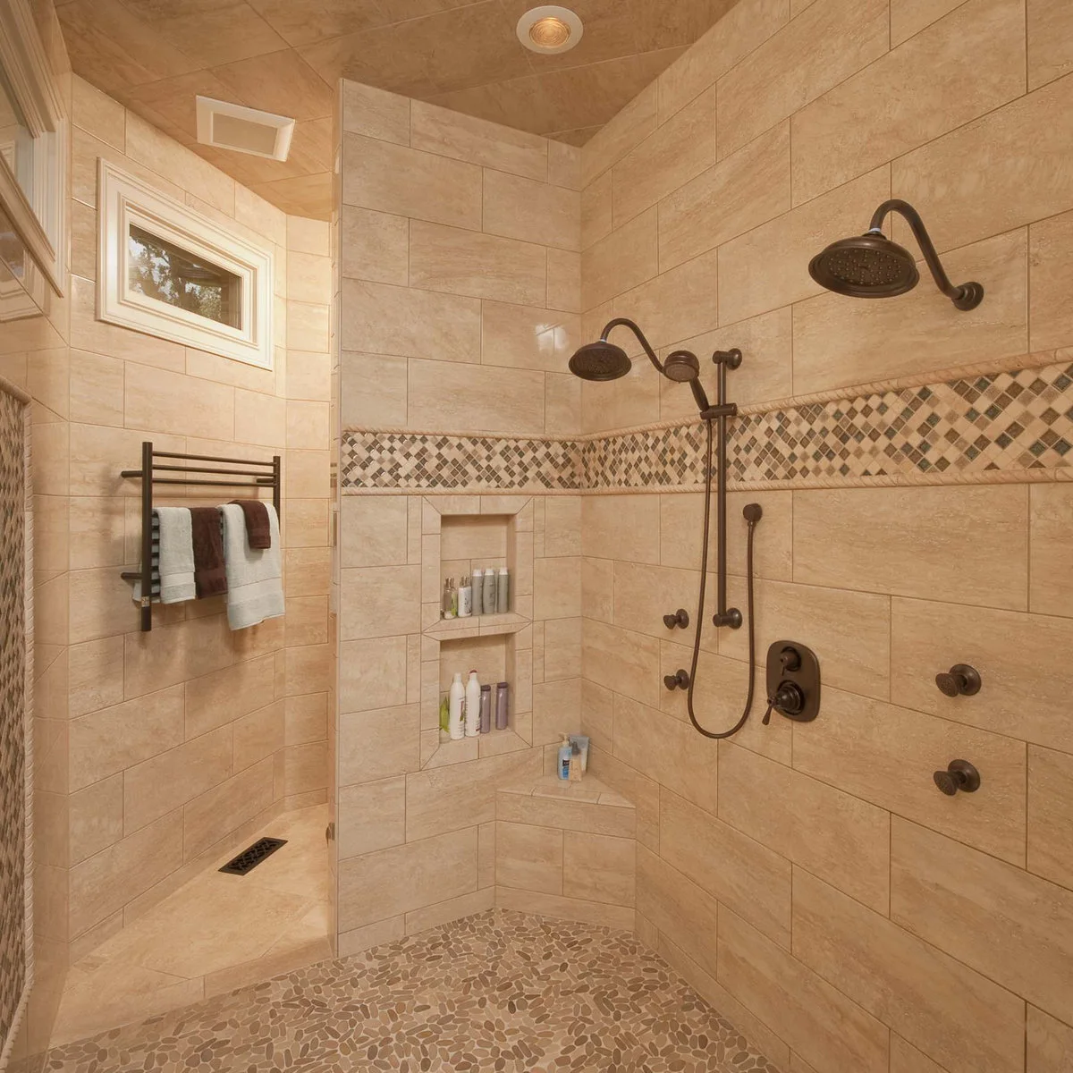 A tiled partition shelters an open shower with two dark showerheads, stacked niches and a pebble-patterned floor.