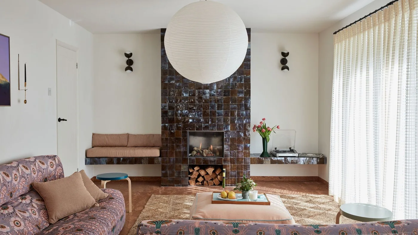 Glossy varied brown square tiles cover a tall fireplace and low side ledges in a room with patterned sofas and a large white pendant.