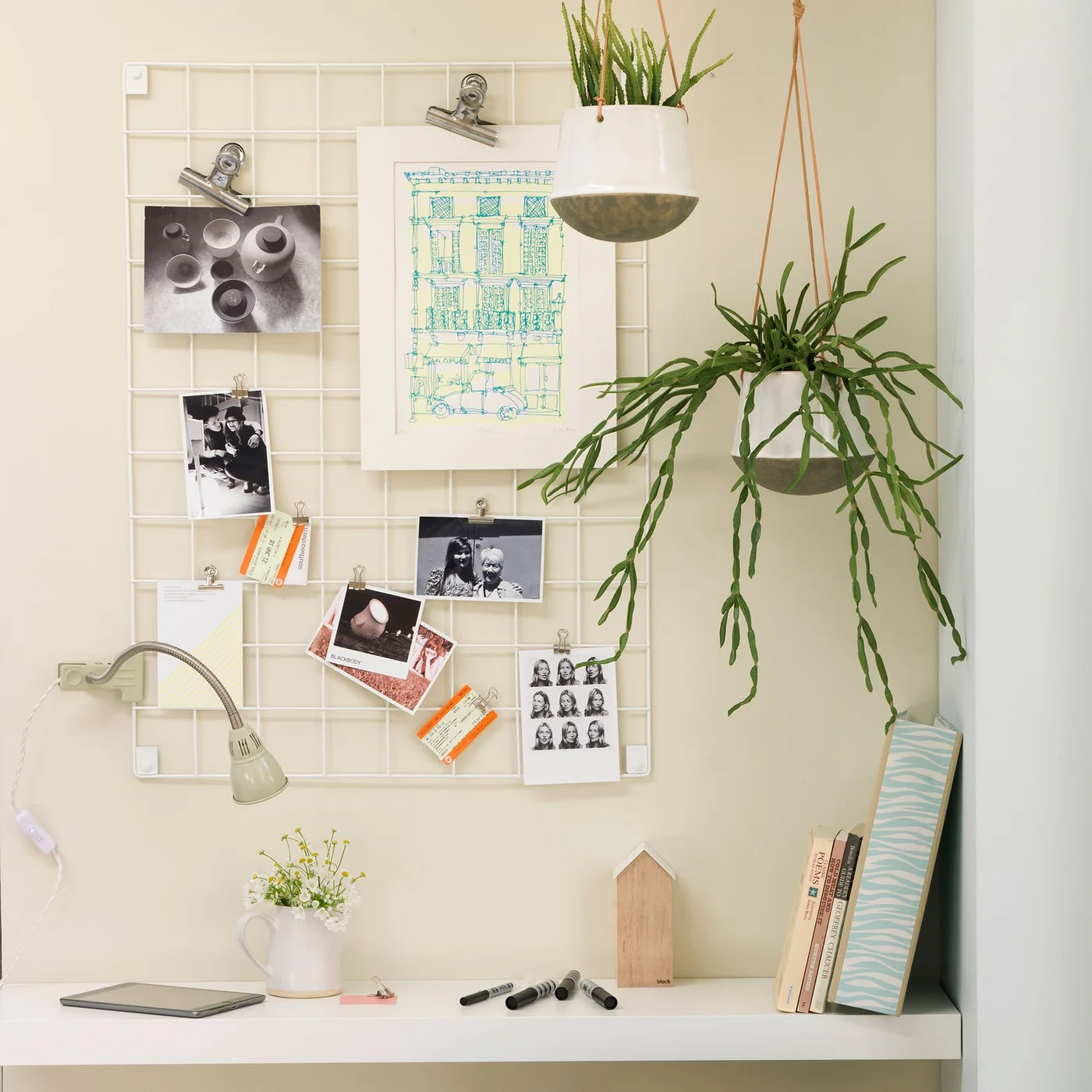 Two white pots hang above a slim white desk beside a wire memo board, with narrow upright and trailing foliage and a small flower jug below.