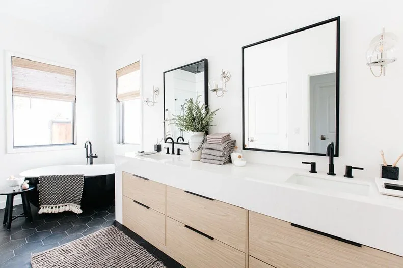 A pale wood double vanity with straight dark handles stands under large dark-framed mirrors beside pale walls, dark taps and hexagonal floor tiles