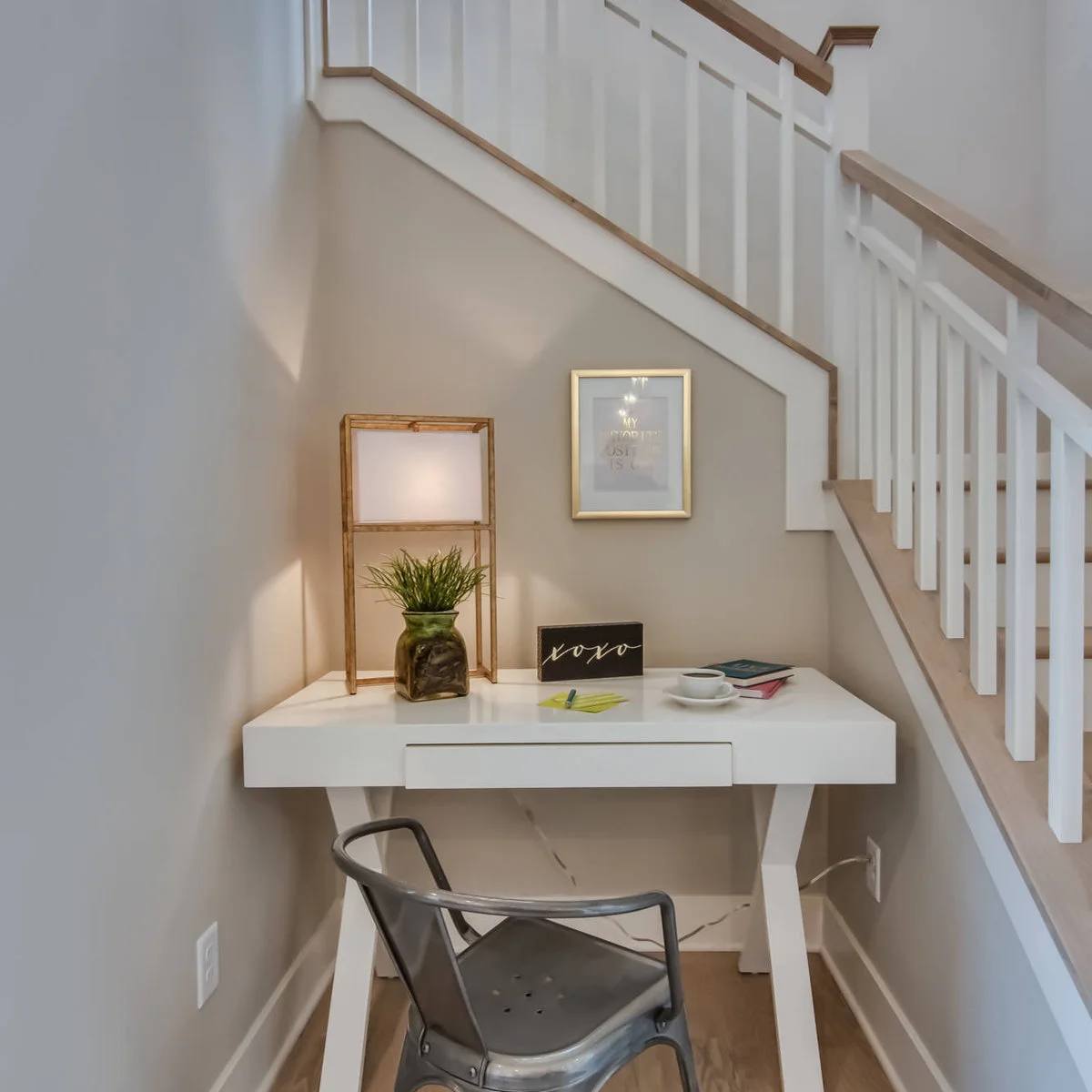 Small white trestle-style desk beneath a staircase with a metal chair and tall rectangular lamp