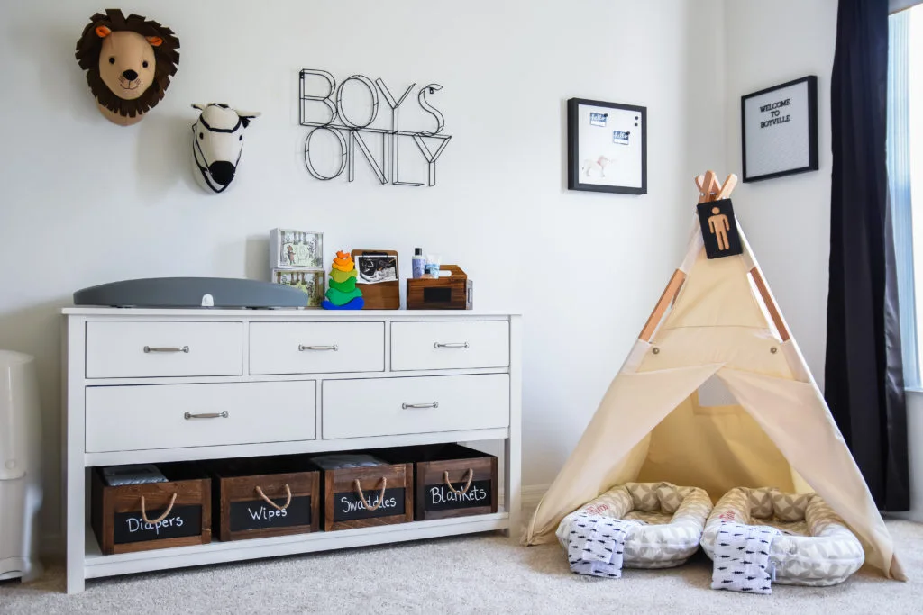 A pale nursery changing cabinet has a gray pad and labeled wood boxes beneath drawers beside a pale play tent and patterned floor props