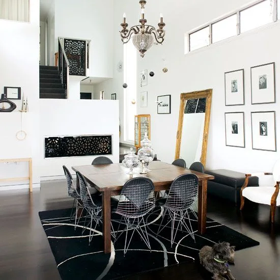 A wood dining table with dark wire-frame chairs stands on a black patterned rug beneath white walls, framed pictures and a chandelier
