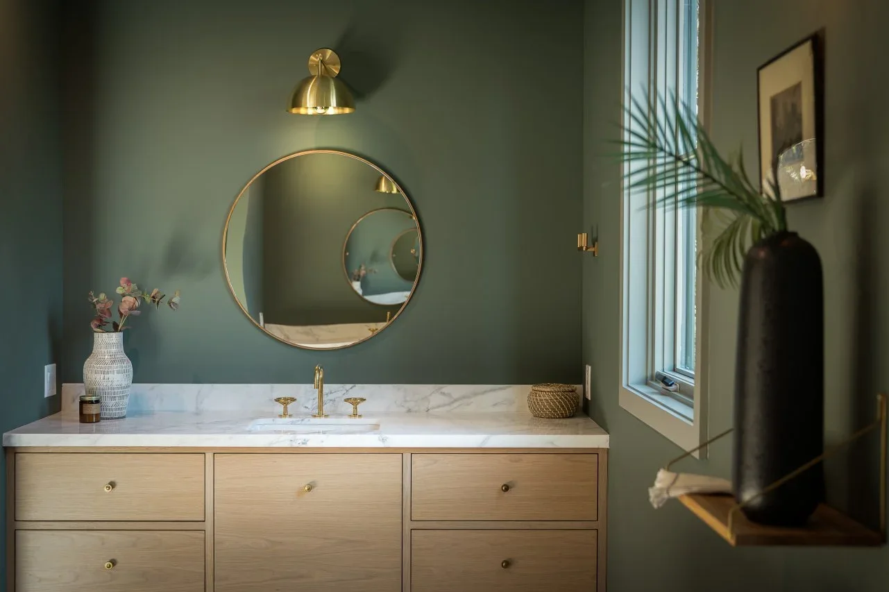 An olive-painted bathroom has a wood drawer vanity, pale countertop, round gold-framed mirror and a window at the side.