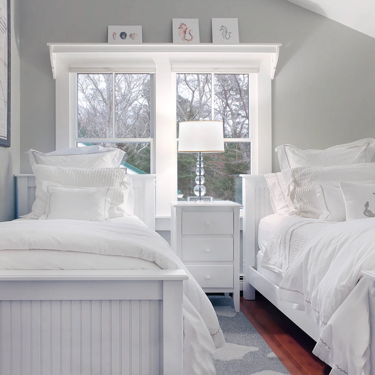 Two white beds with pale bedding and a slim three-drawer chest beneath a double window