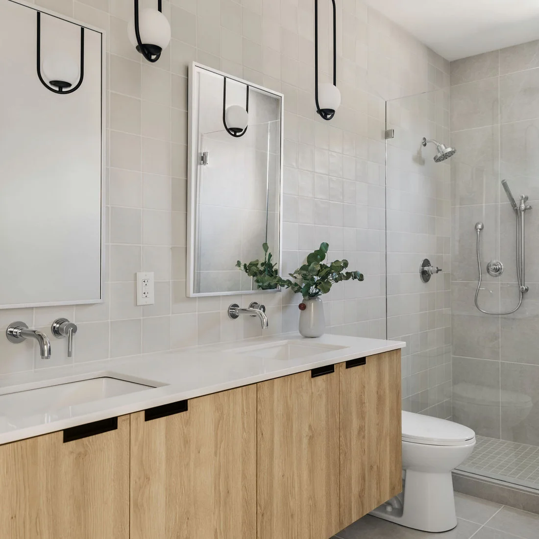 A pale wood double vanity has white-framed mirrored cabinets, chrome wall taps and two black-framed globe pendants, beside a clear shower enclosure and toilet.