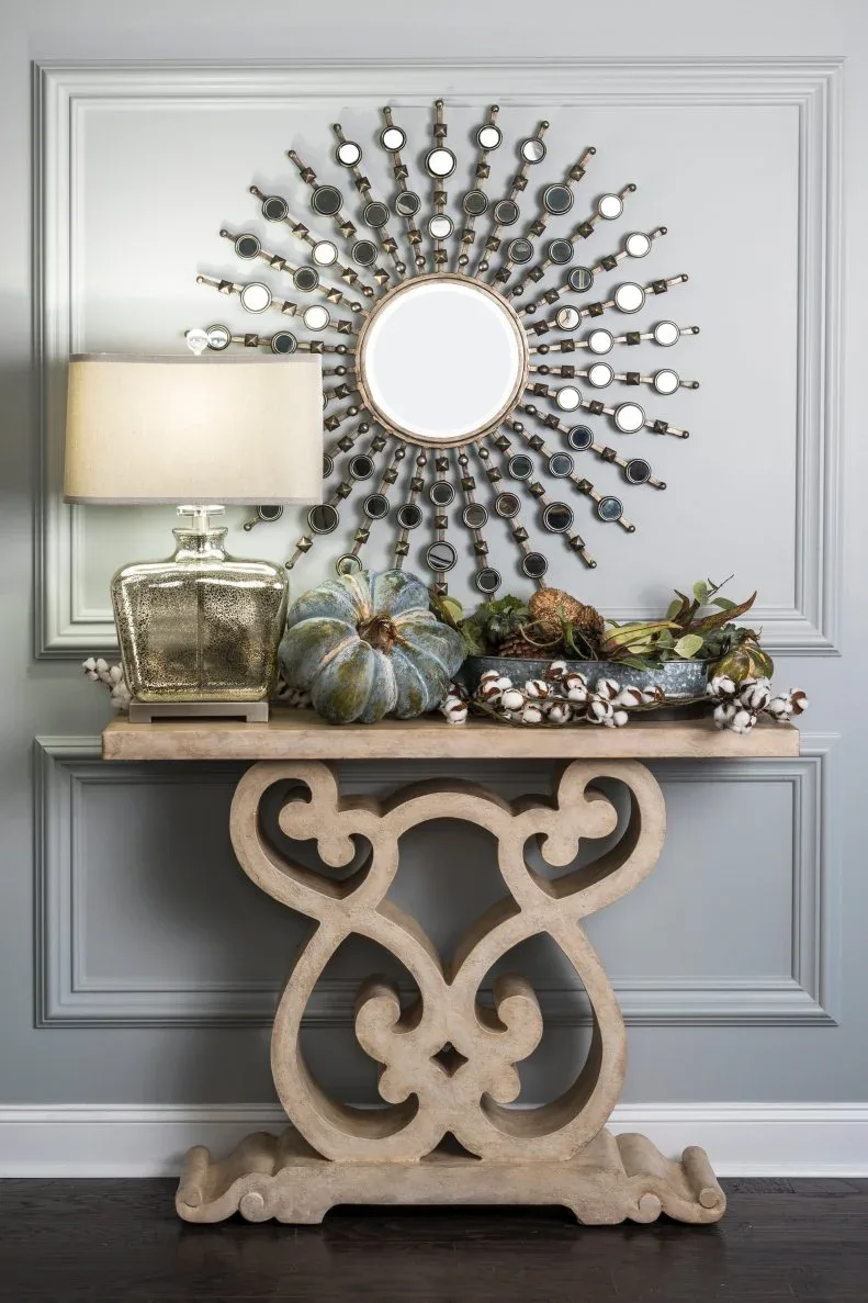 Pale narrow scroll-supported console with blue-gray pumpkins, table lamp and low foliage beneath a radiating mirror